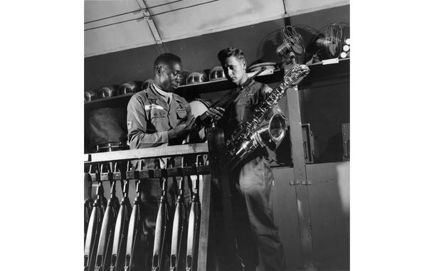 [PUBLISHED CAPTION: Dual mission of the band is shown as SP1 Odis Brown checks a carbine in the supply room before issuing it to SP3 Andrew J. Drazga, who turns in his saxophone. 06/17/1958]; Equally at home on the parade ground or on the maneuvers, members of the 1st Cavalry Division band can switch quickly from one side of their dual role to the other. As bandsmen, the band members provide music for 1st Cavalry activities during normal times, but as soldiers they are ready to exchange their horns and drums for carbines and packs on short notice. When the division moves into the field on maneuvers or in combat, the band becomes a security unit, responsible for perimiter defense of a division command area. [cg]