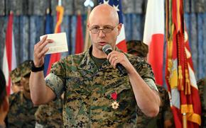 A Marine in camouflage uniform speaks into a microhone held in his left hand while holding up an index card in his right hand and standing in front of U.S. and Japanese flags.