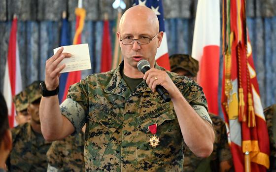 A Marine in camouflage uniform speaks into a microhone held in his left hand while holding up an index card in his right hand and standing in front of U.S. and Japanese flags.