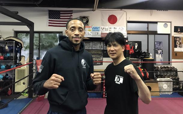 An airman and his trainer both wear workout gear and pose in boxing stances at a gym with American and Japanese flags in the background. 