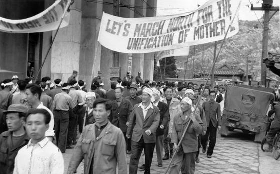 A black-and-white, 1950s-era image of South Korean citizens marching down a street holding a large banner that reads “Let’s march north for the unification of the motherlands.”