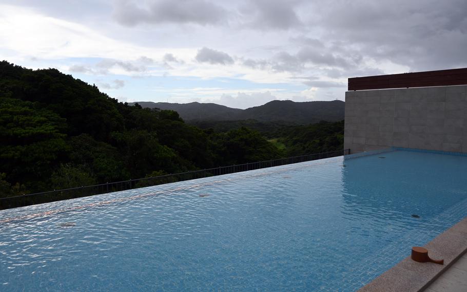 An infinity pool with clear blue water overlooking a lush, forested mountain landscape under a cloudy sky, with modern concrete architecture visible on the right side.