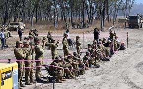 Military personnel in camouflage uniforms gather behind pink safety tape barriers, with some standing and others sitting on the ground. Military vehicles including trucks are visible in the background.