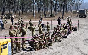Military personnel in camouflage uniforms gather behind pink safety tape barriers, with some standing and others sitting on the ground. Military vehicles including trucks are visible in the background.