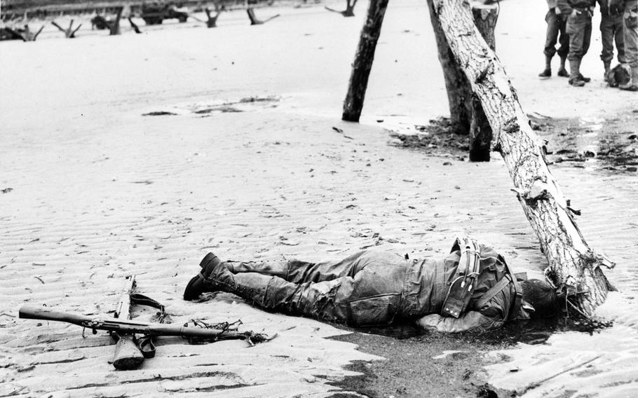 An American Soldier lies dead alongside an anti-landing craft obstruction on Omaha Beach, 6 June 1944. He is wearing an inflatable life belt. Note rifles by his feet, an M1 semiautomatic rifle on the sand, with a M1903 bolt-action rifle laid across it.  