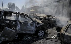 Smoke rises from burned and destroyed cars on a road in front of damaged buildings.