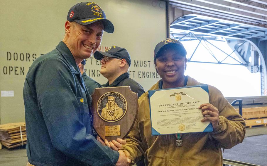 A Navy officer in tan jacket holds up a commendation award while shaking hands and posing with another Navy officer in blue uniform  holding a plaque.
