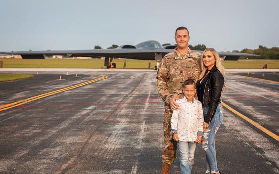 A man in a camouflage uniform poses with a woman and a boy on a military runway with a fighter jet in the background.