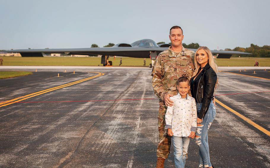 A man in a camouflage uniform poses with a woman and a boy on a military runway with a fighter jet in the background.