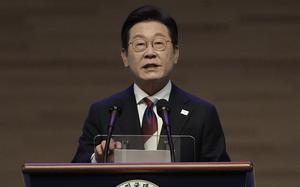 A Korean man wearing a dark suit and glasses stands at a wooden podium and speaks into two microphones in front of a blured brown background.