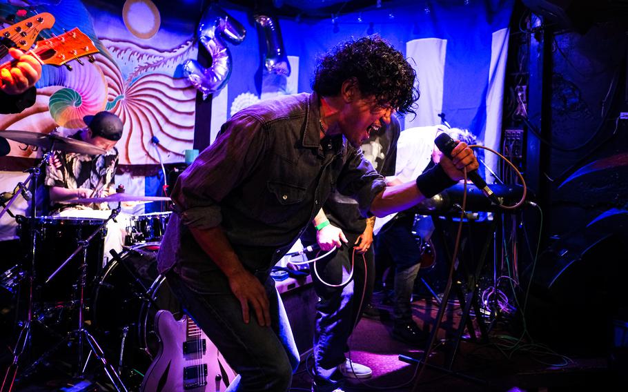 A Japanese man leans forward to sing into a microphone on a stage with a drummer behind him.