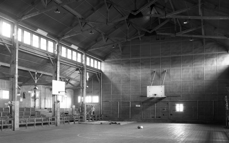 A black and white photo of a gym with a high ceiling, wooden benches serving as bleachers and a basketball hoop.