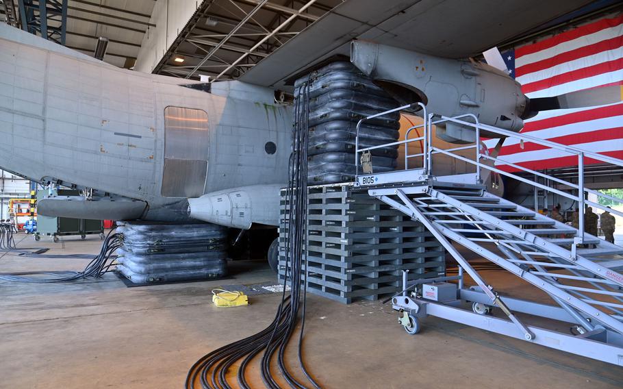 Side view of a military aircraft showing the cargo bay area with the ramp lowered onto multiple layers of black inflatable support bags. Mobile stairs provide access to the aircraft. 