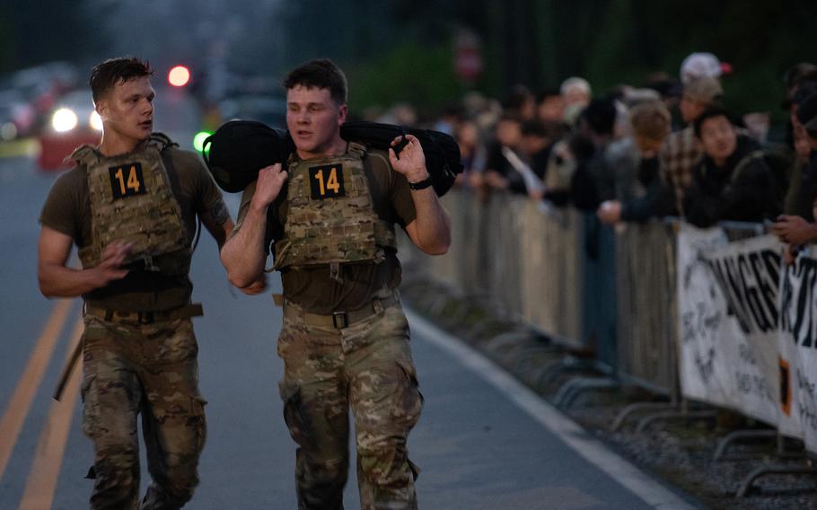 Two soldiers walk down a road as onlookers watch from the sideline, behind a barrier.