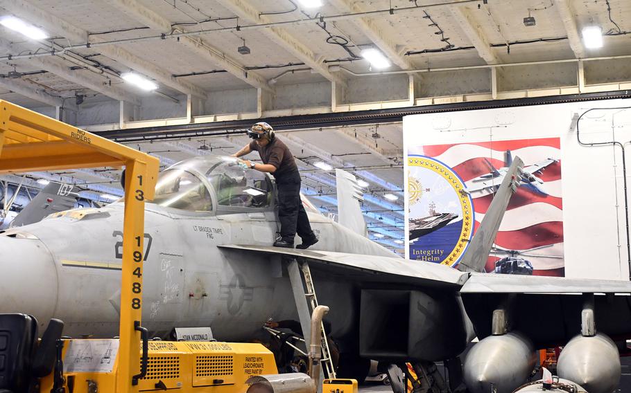A sailor on a ladder works on a fighter jet in a hangar.