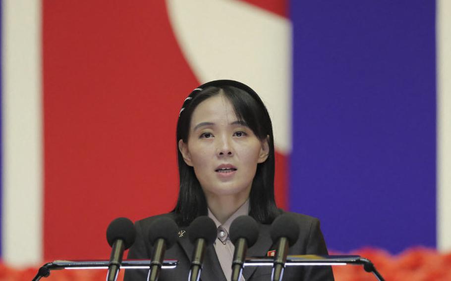 A woman in a dark business suit speaks at a podium with multiple microphones. Red, white and blue colors are visible in the blurred background.