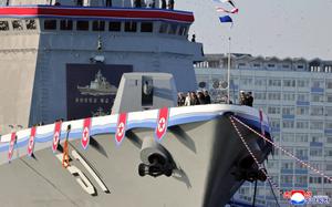 Military officials, seen from a distance, stand at the bow of a warship adorned with North Korean flags down the side.