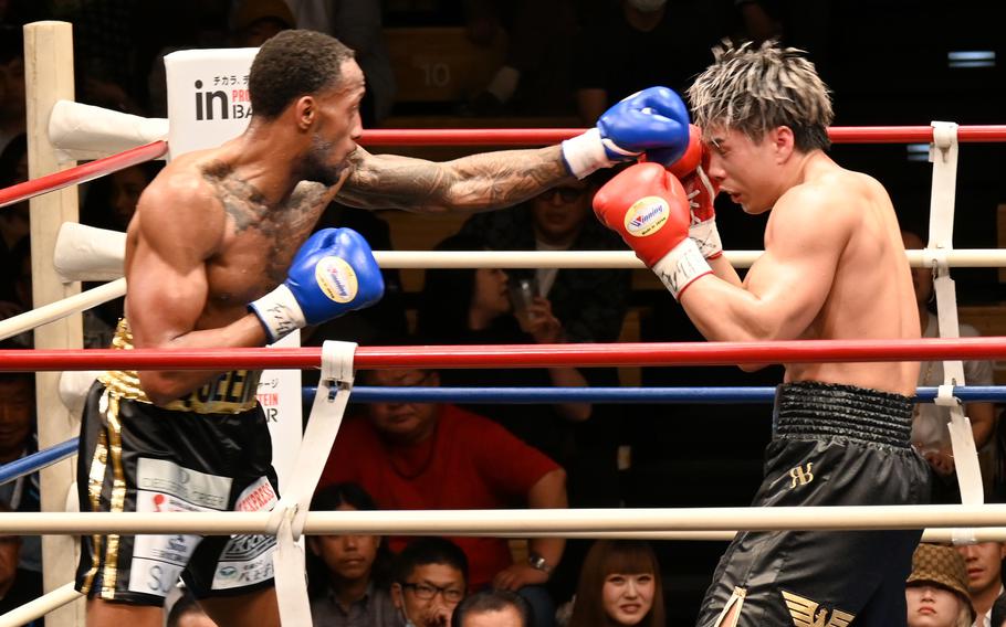 A boxer wearing black shorts and blue gloves throws a punch at the face of another boxer wearing black shorts and red gloves in a ring with a crowd in the background.