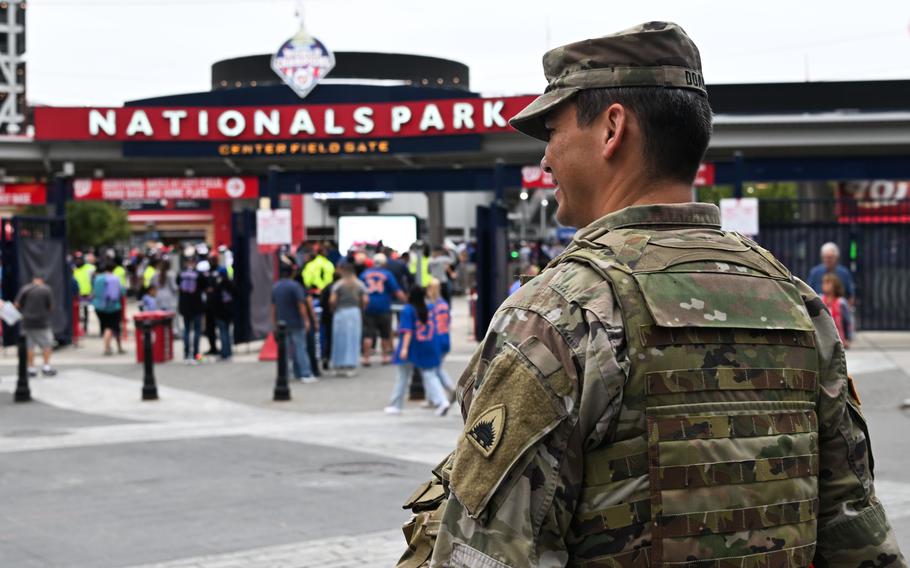 A soldier in camouflage uniform, seen from behind, stands on a concourse outside an entrance gate of a baseball stadium, with a facade reading “Nationals Park.”