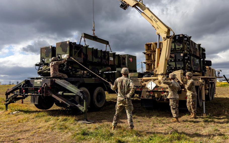 Military personnel in camouflage uniforms operate a yellow crane truck to lift and position green military equipment modules in an open field.