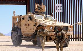 Two soldiers in camouflage combat gear and helmets stand in front of an armored vehicle parked by a section of border wall in desert terrain.