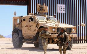 Two soldiers in camouflage combat gear and helmets stand in front of an armored vehicle parked by a section of border wall in desert terrain.