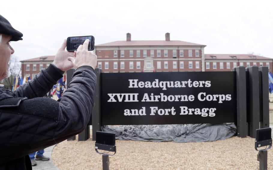 A person in a dark cap and jacket takes a photo with their smartphone of the entrance sign to “Headquarters XVIII Airborne Corps and Fort Bragg,” with red brick military buildings visible in the background.