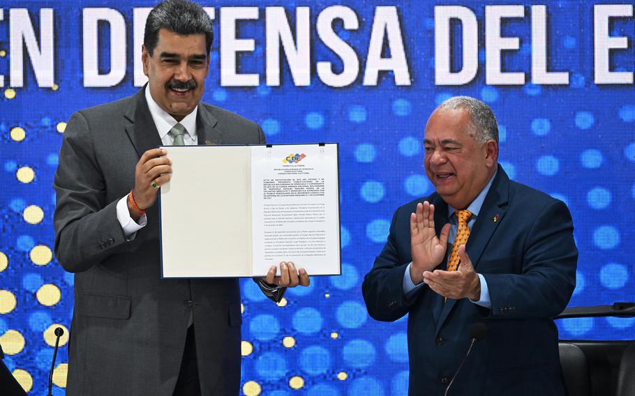 Nicolas Maduro smiles while holding an official document in a folder, as another man next to him smiles and claps.