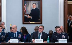 Three men sit at a long desk with placards describing their names and a portrait on the wall behind them.