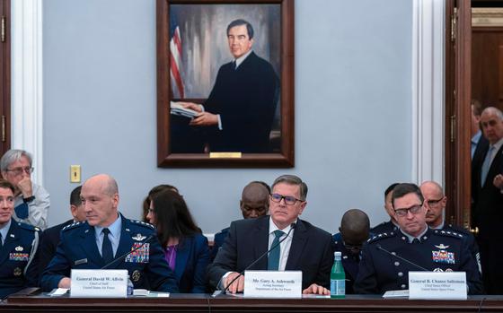 Three men sit at a long desk with placards describing their names and a portrait on the wall behind them.
