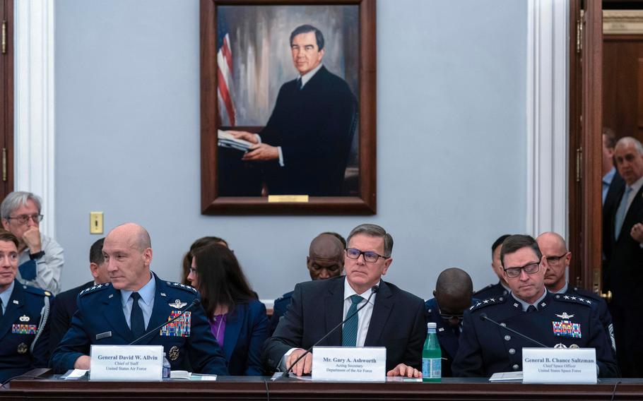 Three men sit at a long desk with placards describing their names and a portrait on the wall behind them.