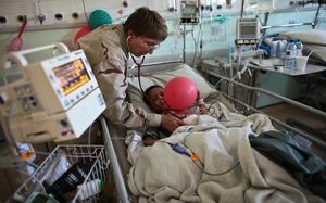 A young boy lies in a hospital bed holding a pink balloon while a doctor leans over him and holds a stethoscope to his chest.