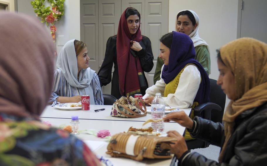 A group of women wearing head scarves speak to each other while standing and sitting around a conference table with plates and drinks on it.
