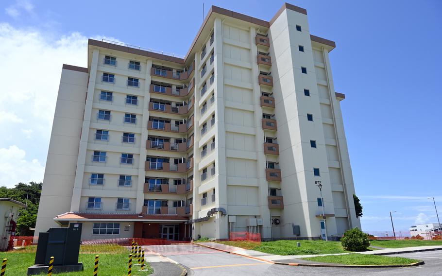 Multi-story residential apartment building with white and beige exterior, featuring multiple balconies and windows on each floor. The building has the number ‘872’ visible on its facade. Orange safety fencing surrounds the perimeter of the property, with a paved driveway leading to the entrance.