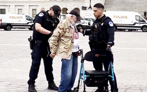 An elderly man in blue jeans, beige jacket and black hat stand between two police officers in uniform as one places zip ties on his wrists and the other holds onto a walker.