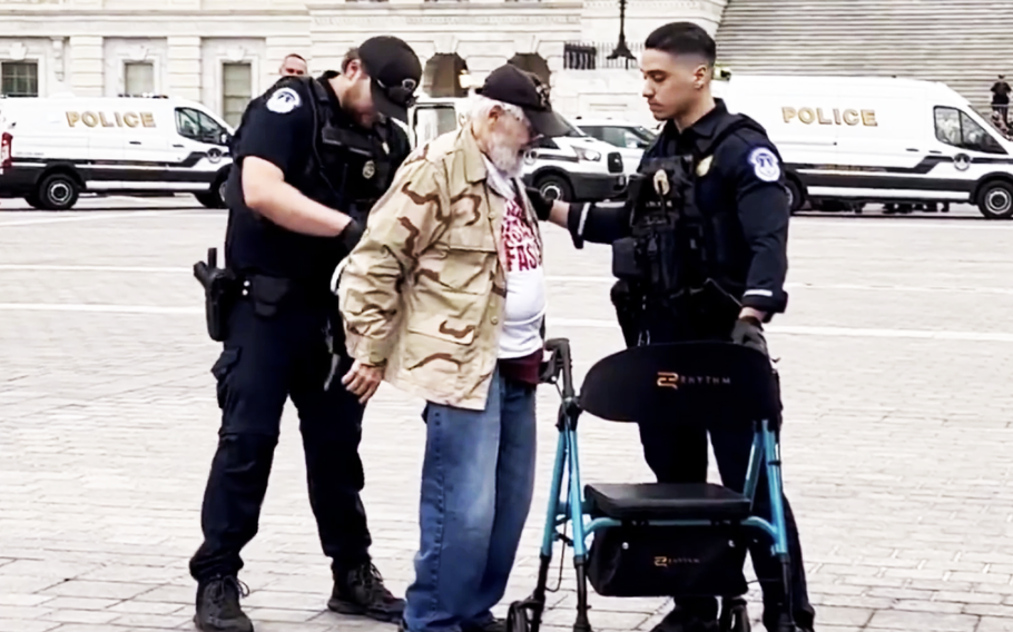 An elderly man in blue jeans, beige jacket and black hat stand between two police officers in uniform as one places zip ties on his wrists and the other holds onto a walker.