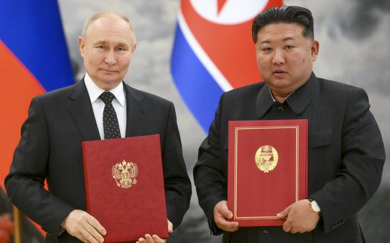 Vladimir Putin and Kim Jong Un pose next to each other while holding red folders with gold emblems in front of Russian and North Korean flags.