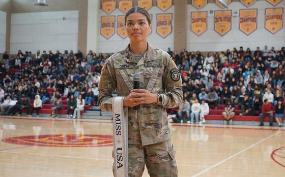 A soldier with a Miss USA sash stands in front of a crowd of students in a gymnasium holding a microphone