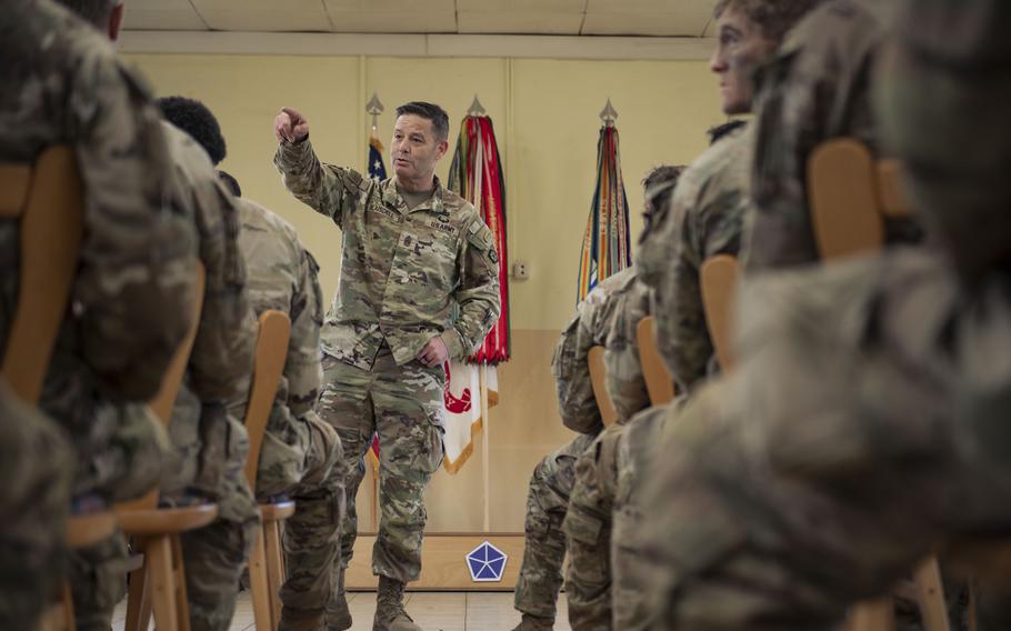 The legs and backs of soldiers seated are seen as they listen to a Command Sgt. Mag.