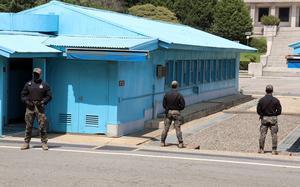 Three soldiers in camouflage uniforms and black jackets stand guard near distinctive blue military buildings at the DMZ with corrugated metal roofs. 