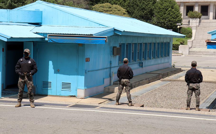 Three soldiers in camouflage uniforms and black jackets stand guard near distinctive blue military buildings at the DMZ with corrugated metal roofs. 