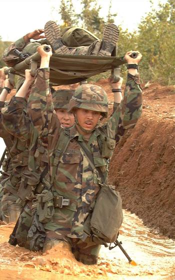 A line of soldiers hoisting another soldier on a stretcher above their heads; all are wading through muddy water.