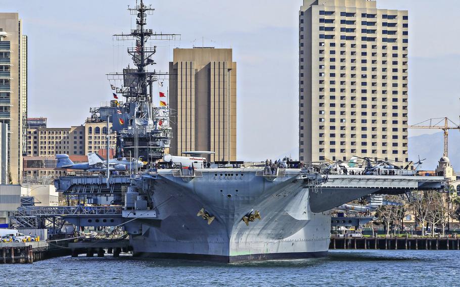 An aircraft carrier, seen from the front, sits docked at a pier with a city skyline in the background.