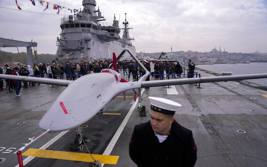 A sailor stands on deck with a drone and a crowd of people in the background.