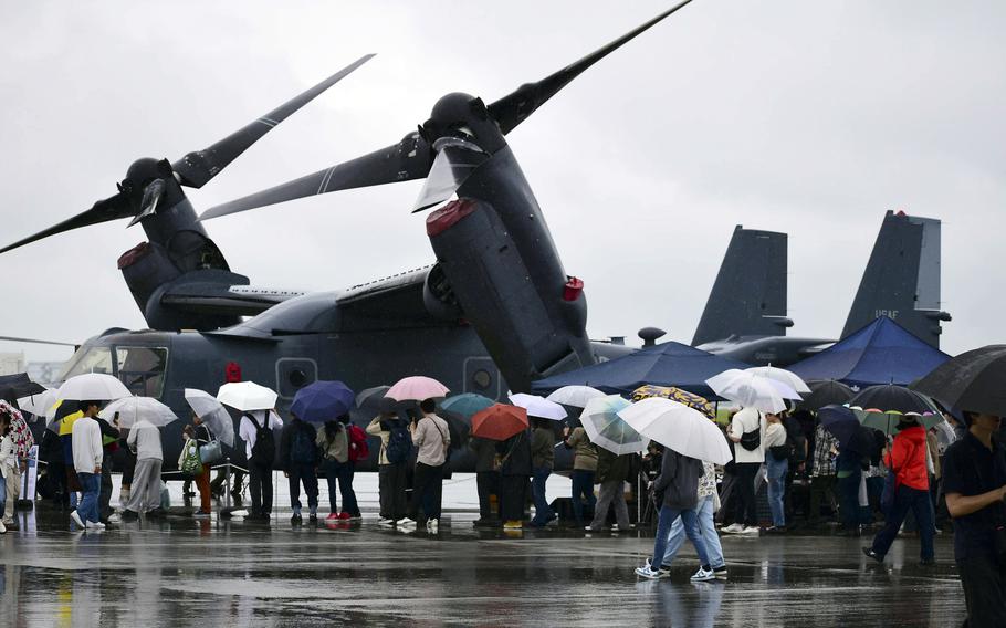 People with umbrellas line up on a rainy day with a CV-22 Osprey in the background.
