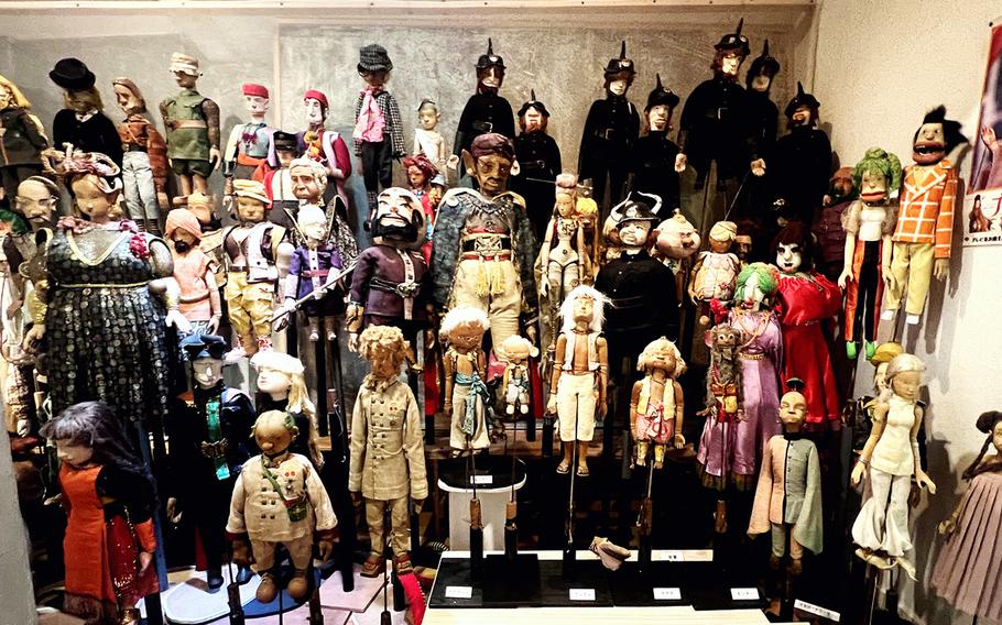 Several dozen puppets sit in rows in a display in front of a white wall.