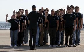 A group of Air Force recruits stand in rows on a runway in front of an officer in uniform with their right hands raised to take their enlistment oath.