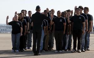 A group of Air Force recruits stand in rows on a runway in front of an officer in uniform with their right hands raised to take their enlistment oath.