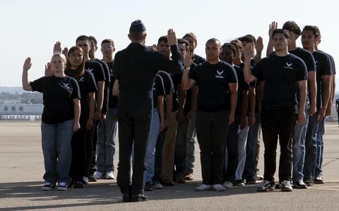A group of Air Force recruits stand in rows on a runway in front of an officer in uniform with their right hands raised to take their enlistment oath.