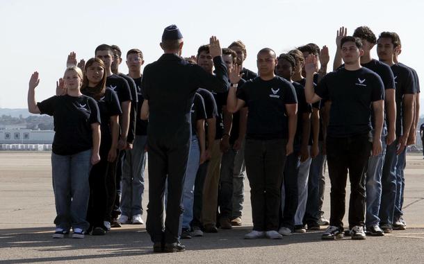A group of Air Force recruits stand in rows on a runway in front of an officer in uniform with their right hands raised to take their enlistment oath.